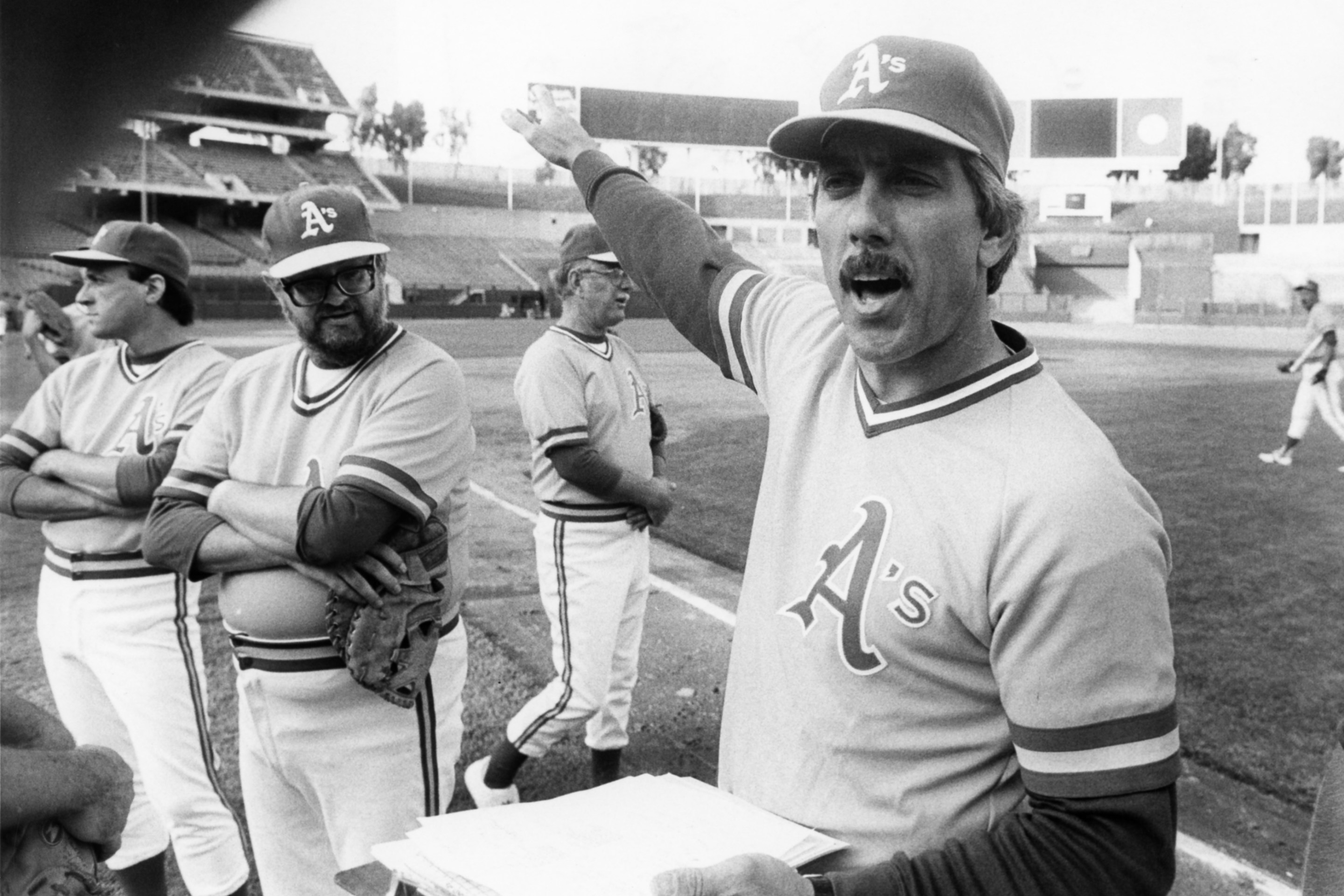 A black-and-white image of Ray Fosse, in an old-timey Oakland A's uniform, bossing around a bunch of lumpy '70s guys at an A's fantasy camp in