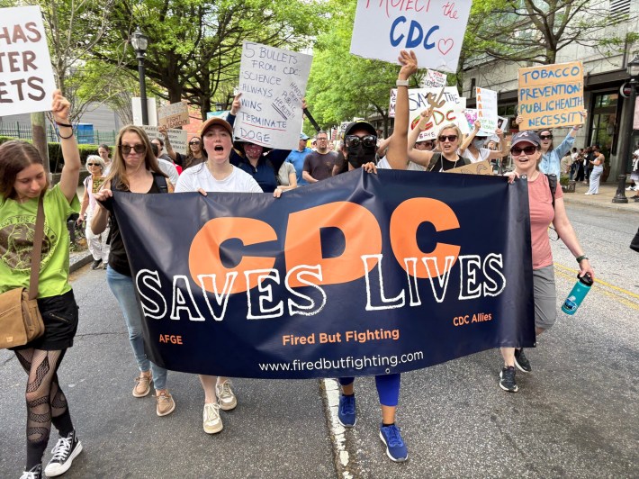 A photo of a group of marching protesters carrying a banner saying CDC SAVES LIVES, AFGE, Fired But Fighting, CDC Allies, and www.firedbutfighting.com