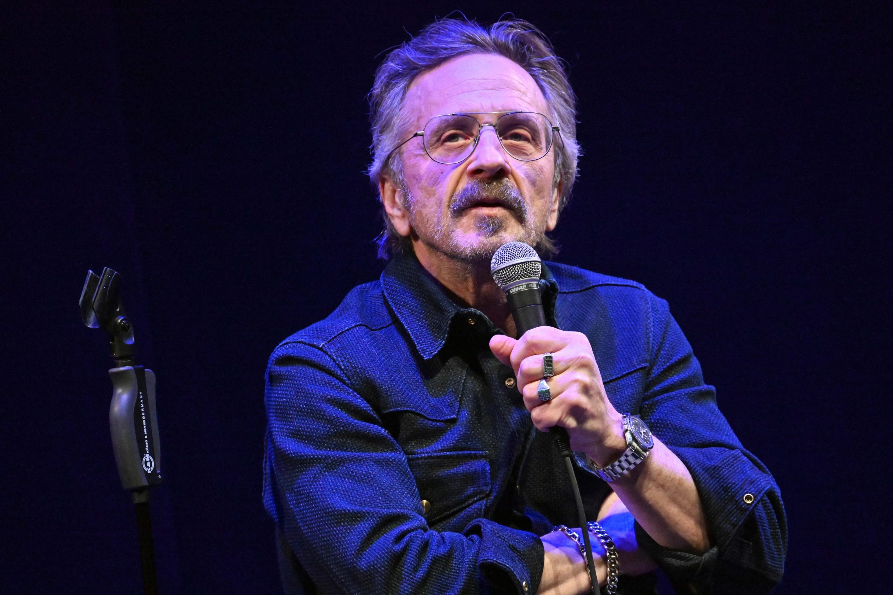 Marc Maron performing stand up comedy. He is sitting on a stool and holding a microphone